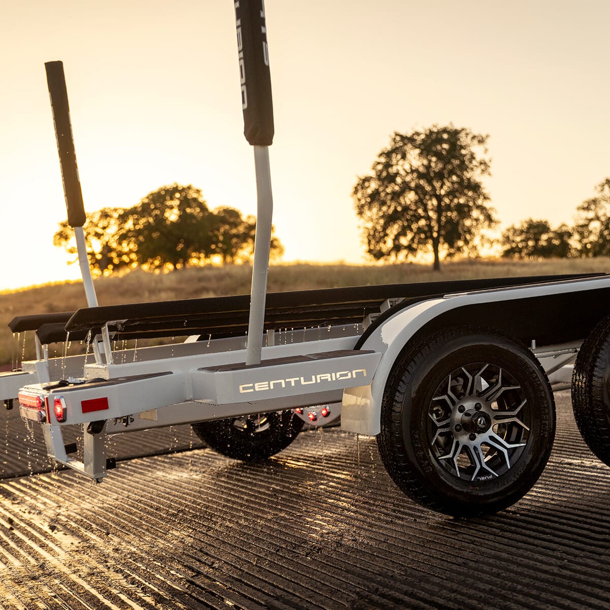 A white Centurion trailer with double axles sits on a grooved ramp at sunset, with trees visible in the background.