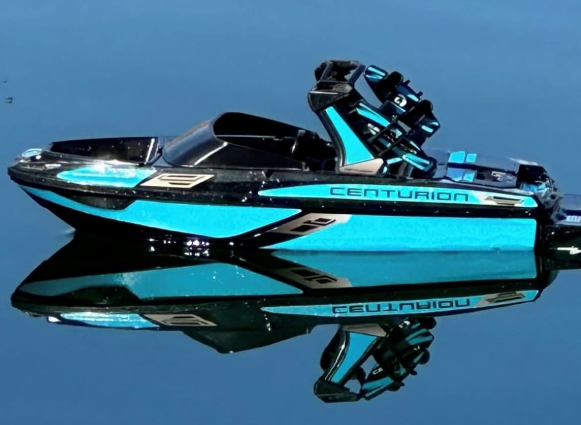 A blue Centurion wakeboard boat floats on calm water, perfectly reflecting its image.