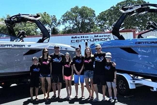 A group of nine people, including adults and children, stand in front of two parked Centurion boats outside a Mello Marine dealership. They are all wearing matching T-shirts.