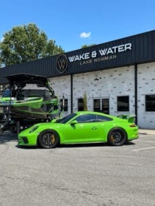 A bright green sports car and a matching green and black boat are parked in front of the Wake & Water Lake Norman building on a sunny day.