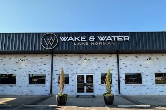 Storefront of Wake & Water Lake Norman with a black and white exterior brick wall, double glass doors, two planter pots with trees on either side, and a clear blue sky.