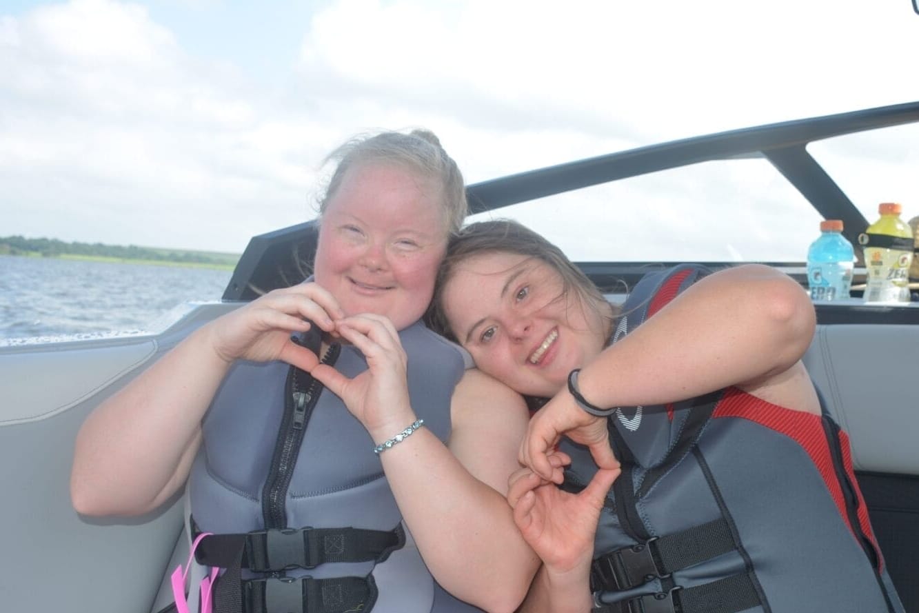 Two people wearing life jackets sit on a boat, making heart shapes with their hands. A third person stands nearby, and drinks are visible in the background.