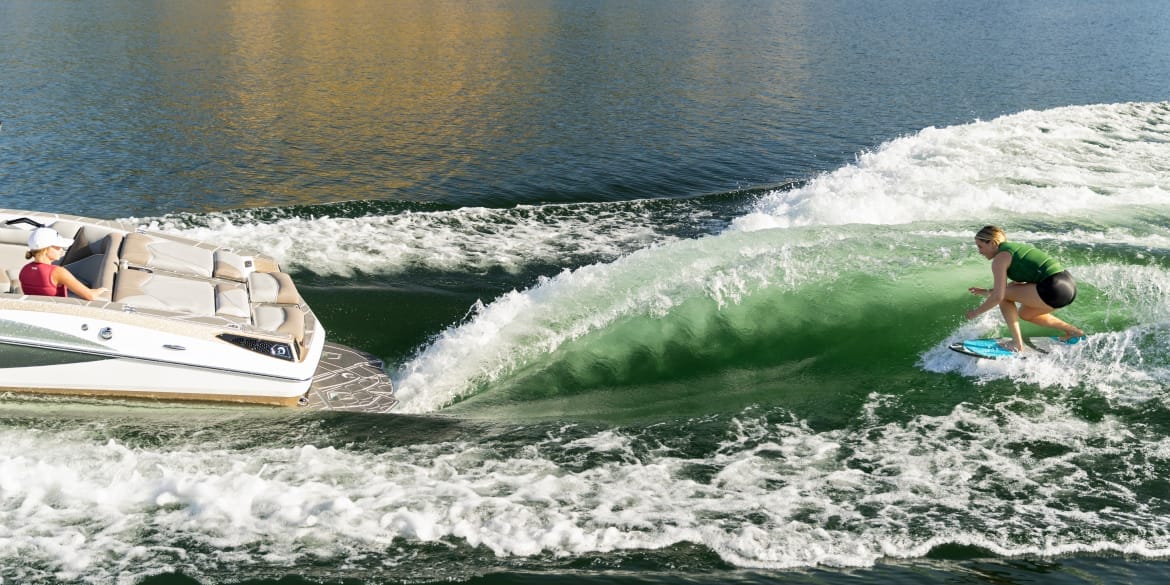 A person surfs on the wave created by a boat, with one person sitting at the back of the boat on a sunny day. Discover Centurion Boats and experience unparalleled water adventures.
