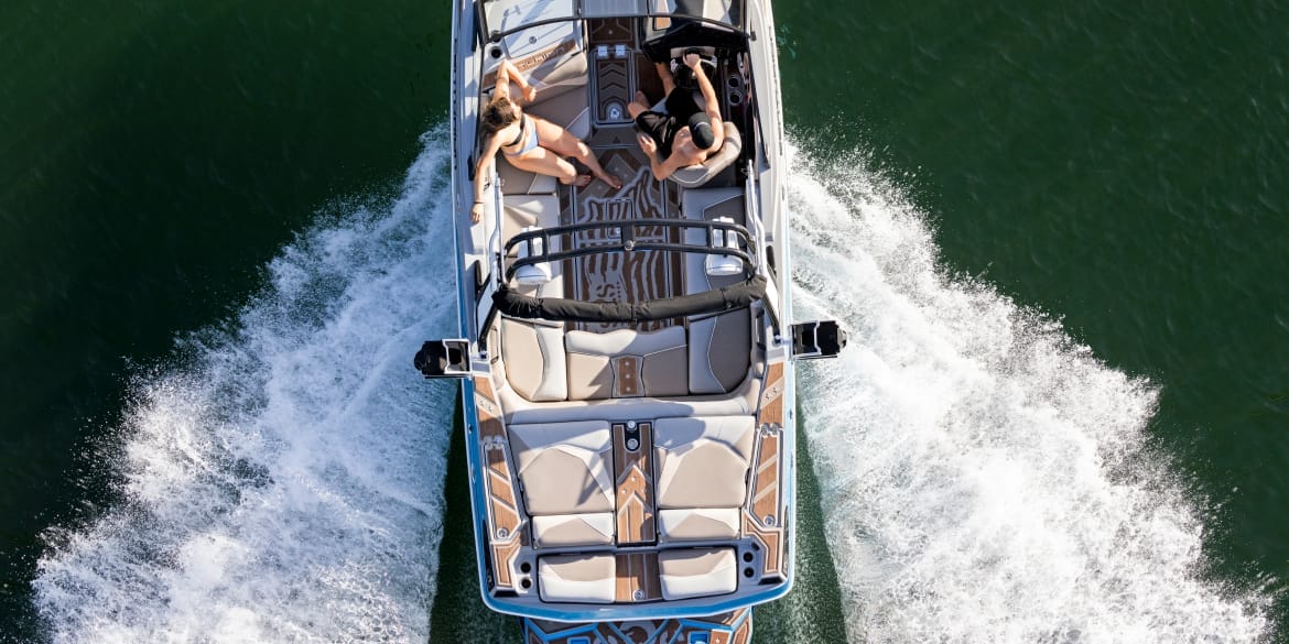 Aerial view of a speedboat on a body of water with two women sunbathing on the deck. Discover Centurion Boats as it leaves a wake in the water, moving forward gracefully.