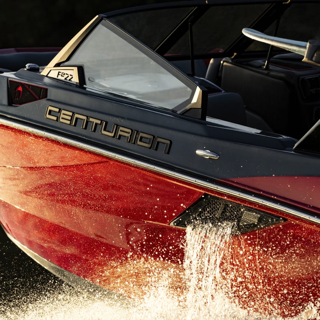Close-up of a red and black Centurion boat cutting through water, creating a splash. The boat is viewed from the side, showcasing its sleek design.