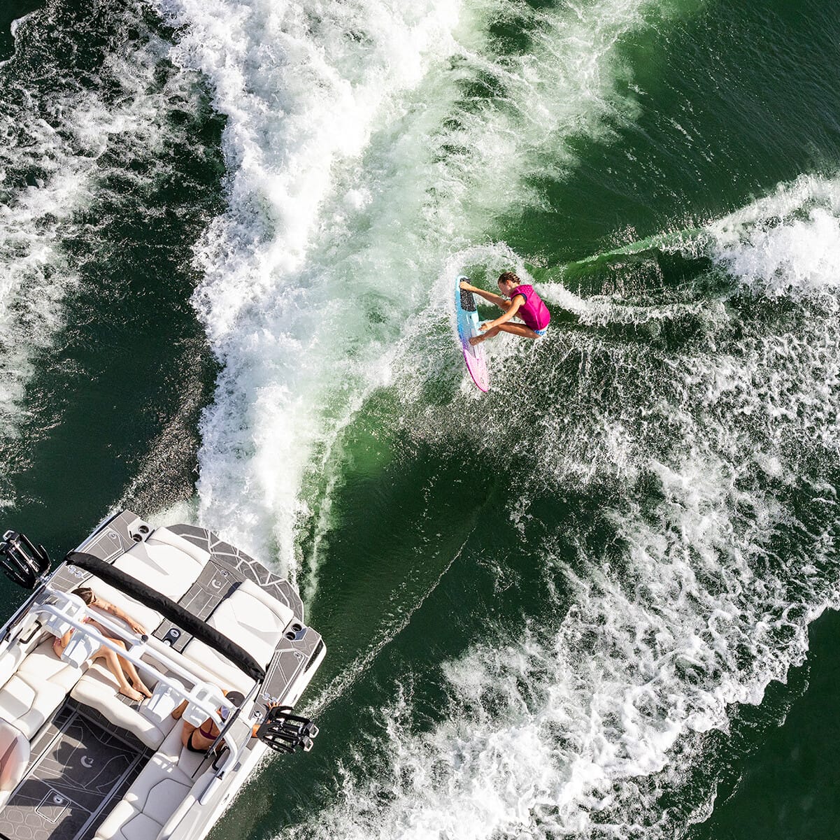A person surfs on a wakeboard behind a motorboat on a lake, creating splash and waves, while three people relax on the boat.