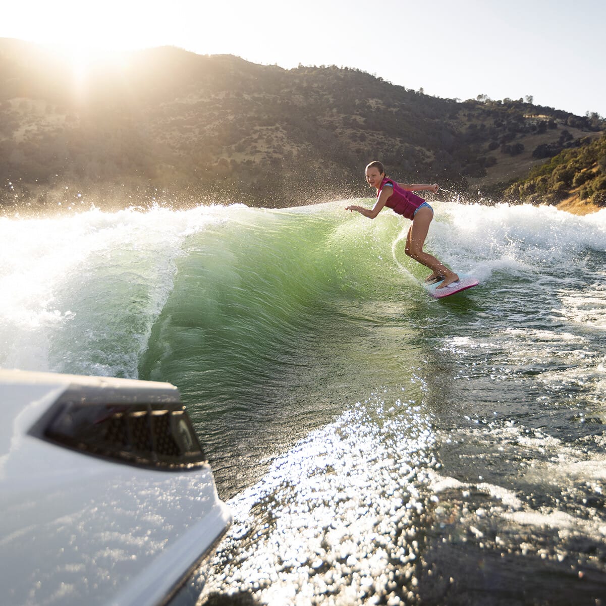 A person surfs on a green wave near a boat in a sunny, mountainous area.
