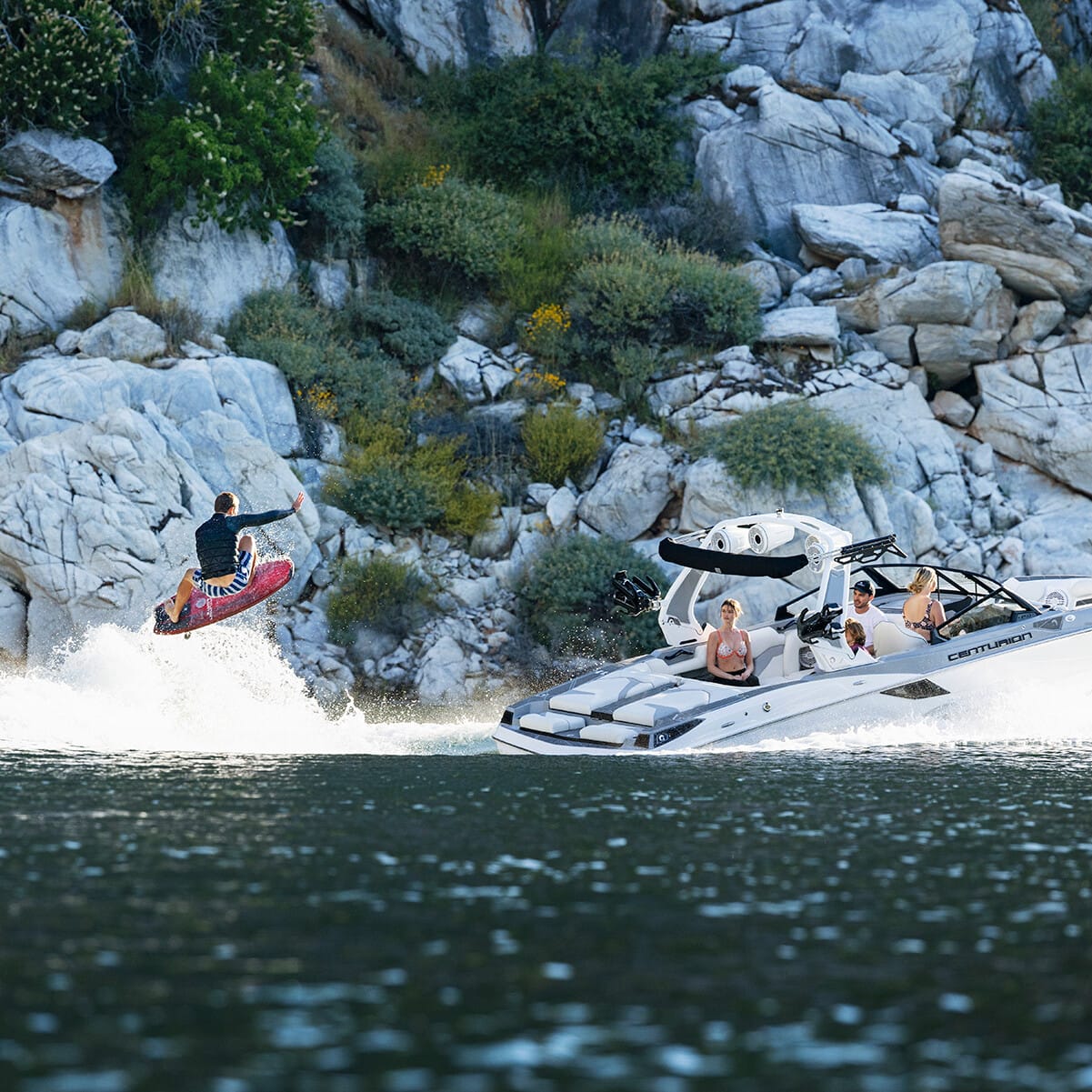 A person is wakesurfing behind a white speedboat with three passengers on a lake surrounded by rocky cliffs and greenery.