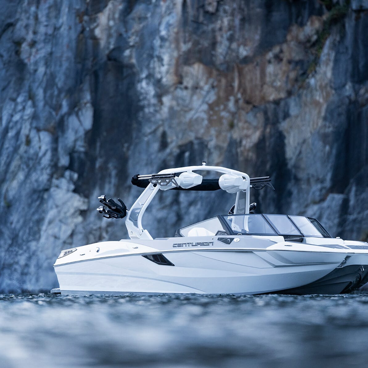 A white speedboat with a wakeboard tower floats on calm water near a rocky cliff.