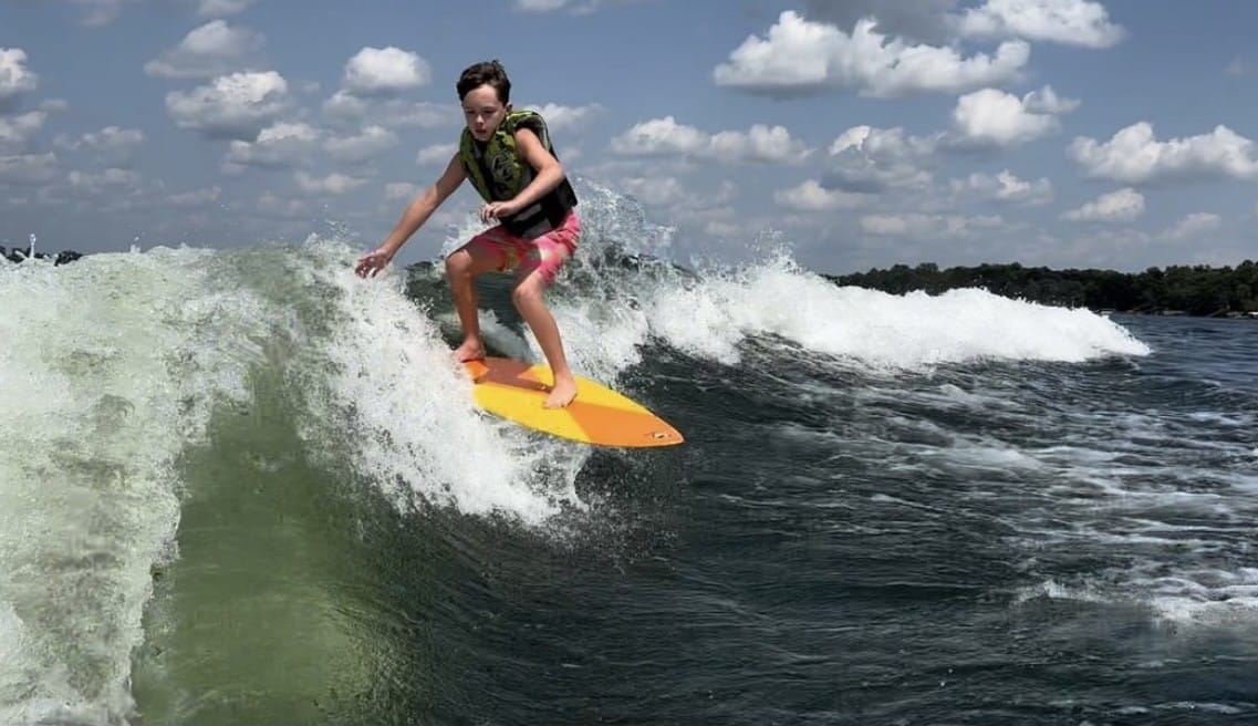 A person wearing a life vest rides a yellow surfboard on a wave under a blue sky with scattered clouds.